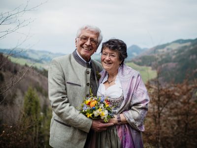 Ein älteres Paar steht lächelnd auf einem Hügel und hält einen bunten Blumenstrauß. Der Mann trägt eine Brille und einen Anzug, während die Frau ein traditionelles Kleid und einen Schal trägt. Hinter ihnen stehen Bäume und Berge unter einem bewölkten Himmel.