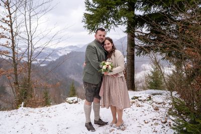 Ein Paar steht lächelnd auf einem verschneiten Berg, der Mann trägt traditionelle Kleidung und die Frau hält einen Blumenstrauß.