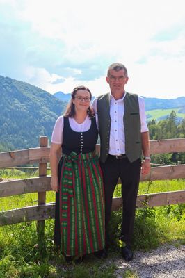 Ein Mann und eine Frau in traditioneller Kleidung stehen nebeneinander vor einem Holzzaun mit einer Berglandschaft im Hintergrund.