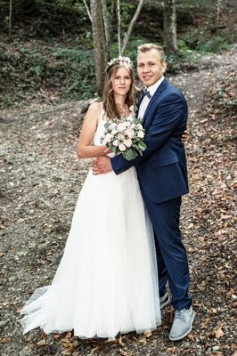 Ein frisch verheiratetes Paar steht eng zusammen auf einem Waldweg, die Braut in einem weißen Kleid und der Bräutigam in einem blauen Anzug, beide lächeln.