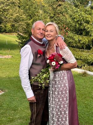 Ein älteres Paar steht gemeinsam in einem Garten, der Mann mit einem Blumenstrauß und einer roten Rose auf seiner Weste, lächelt in die Kamera.