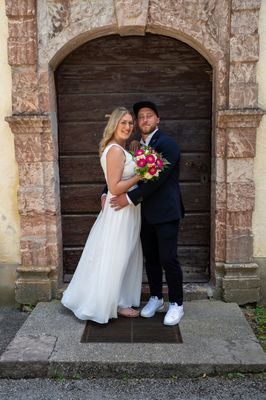 Ein frisch verheiratetes Paar steht vor einer alten Holztür, die Braut in einem weißen Kleid und der Bräutigam in einem dunklen Anzug, beide lächeln und halten einen Blumenstrauß.