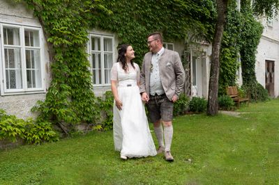 Ein frisch verheiratetes Paar steht vor einem mit Efeu bewachsenen Haus, die Braut in einem weißen Kleid und der Bräutigam in traditioneller deutscher Kleidung.