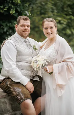 Ein Mann und eine Frau in traditioneller deutscher Kleidung lächeln und posieren für ein Hochzeitsfoto. Der Mann trägt eine Weste und die Frau hält einen Blumenstrauß.