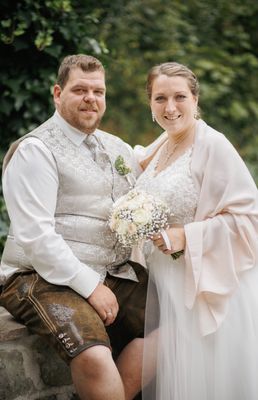 Ein Mann und eine Frau in traditioneller deutscher Kleidung lächeln und posieren für ein Hochzeitsfoto. Der Mann trägt eine Weste und die Frau hält einen Blumenstrauß.