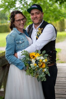 Ein lächelndes Paar, die Frau in einem weißen Kleid und einer Jeansjacke, der Mann in einem schwarzen Weste, stehen zusammen auf einer Holzbrücke mit einem Strauß Sonnenblumen und Grün.