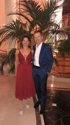 Ein Mann und eine Frau stehen lächelnd eng beieinander in einem gut beleuchteten Innenbereich mit einer großen Palme und einem dekorativen Geländer hinter ihnen.