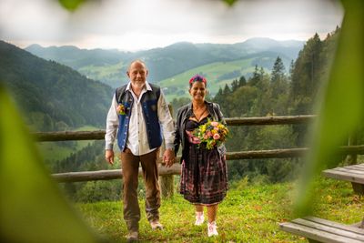 Ein Paar steht auf einem Grashügel, Händchen haltend, mit einem Holzzaun und einer Berglandschaft im Hintergrund. Der Mann trägt eine Weste und die Frau hält einen Blumenstrauß.