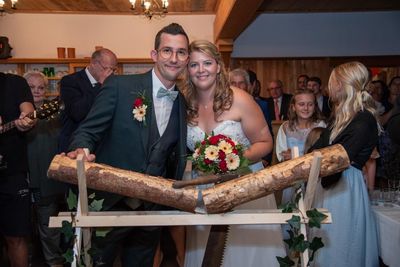 Ein frisch verheiratetes Paar steht hinter einem hölzernen Bogen und lächelt für ein Foto. Der Bräutigam trägt einen Anzug und eine Fliege, während die Braut einen Blumenstrauß hält. Hinter ihnen beobachten die Gäste die Zeremonie.