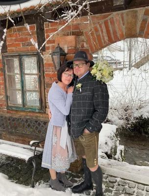 Ein Paar steht eng beieinander in einer verschneiten Umgebung, der Mann trägt einen Hut und einen Blumenstrauß, und die Frau ein hellblaues Kleid. Hinter ihnen steht ein Backsteingebäude mit einem Fenster und einer mit Schnee bedeckten Bank.