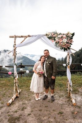 Ein Paar steht unter einem rustikalen Bogen mit Blumendekorationen vor einer Bergkulisse. Die Braut hält einen Blumenstrauß, und der Bräutigam trägt traditionelle Kleidung.