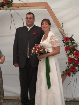 Ein frisch verheiratetes Paar steht zusammen, der Bräutigam in einem schwarzen Anzug und die Braut in einem weißen Hochzeitskleid, hält einen Blumenstrauß. Sie lächeln und scheinen für ein Foto zu posieren.