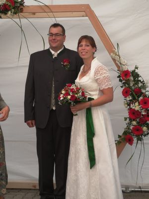 Ein frisch verheiratetes Paar steht zusammen, der Bräutigam in einem schwarzen Anzug und die Braut in einem weißen Hochzeitskleid, hält einen Blumenstrauß. Sie lächeln und scheinen für ein Foto zu posieren.