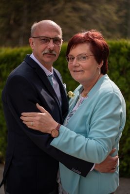 Ein älteres Paar, der Mann in einem dunklen Anzug und die Frau in einem hellblauen Blazer, umarmen sich in einem Garten.