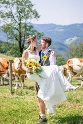 Ein frisch verheiratetes Paar steht in einem Feld mit Kühen im Hintergrund, der Bräutigam hebt die Braut hoch, während sie sich beide anlächeln.