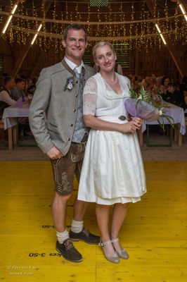 Ein Mann und eine Frau in traditioneller bayerischer Kleidung stehen lächelnd für ein Foto bei einer Hochzeitsfeier. Die Frau hält einen Blumenstrauß.