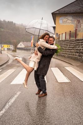 Ein Paar umarmt sich unter einem durchsichtigen Regenschirm bei Regen, mit einer Steinmauer und einer Fahne im Hintergrund.
