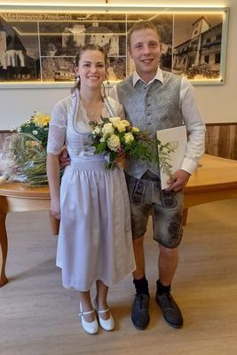 Ein junges Paar in traditioneller Kleidung, lächelnd und posiert für ein Foto mit einem Blumenstrauß und einem Zertifikat in der Hand.