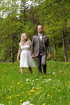 Ein Paar in formeller Kleidung geht Hand in Hand durch ein üppiges grünes Feld mit Bäumen und Blumen im Hintergrund.