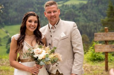 Ein frisch verheiratetes Paar steht lächelnd eng beieinander, die Braut hält einen Blumenstrauß und der Bräutigam trägt einen beigen Anzug vor einem Hintergrund aus üppigen grünen Hügeln.