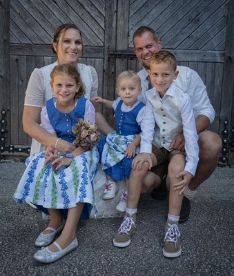 Eine Familie mit vier Personen, darunter zwei Kinder, in traditioneller Kleidung, lächelt für ein Foto vor einer Holzscheune.