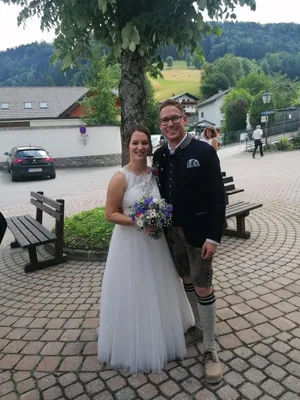 Ein frisch verheiratetes Paar steht lächelnd vor einem Baum, die Frau in einem weißen Hochzeitskleid und der Mann in traditioneller Kleidung. Hinter ihnen ist ein Auto geparkt, und eine Person geht auf dem Bürgersteig.
