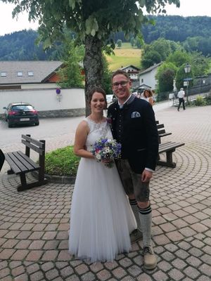 Ein frisch verheiratetes Paar steht lächelnd vor einem Baum, die Frau in einem weißen Hochzeitskleid und der Mann in traditioneller Kleidung. Hinter ihnen ist ein Auto geparkt, und eine Person geht auf dem Bürgersteig.