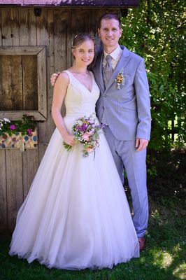 Eine Braut in einem weißen Kleid und ein Bräutigam in einem grauen Anzug stehen zusammen draußen in der Nähe einer Holzwand.