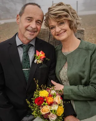 Ein Mann und eine Frau stehen eng beieinander und lächeln für ein Foto. Der Mann trägt einen schwarzen Anzug mit einer grünen Krawatte und einer Blume an seiner Revers. Die Frau trägt eine grüne Jacke und eine Brille.