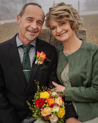Ein Mann und eine Frau stehen eng beieinander und lächeln für ein Foto. Der Mann trägt einen schwarzen Anzug mit einer grünen Krawatte und einer Blume an seiner Revers. Die Frau trägt eine grüne Jacke und eine Brille.