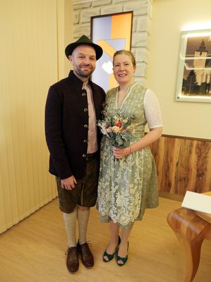 Ein Mann und eine Frau in traditioneller bayerischer Kleidung stehen lächelnd nebeneinander, mit einem Blumenstrauß in den Händen der Frau. Sie befinden sich in einem Raum mit Holzwänden und einem Bilderrahmen an der Wand.