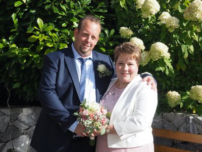 Ein Mann und eine Frau, wahrscheinlich ein Paar, stehen eng beieinander vor einem Busch mit weißen Blumen und lächeln für ein Foto.