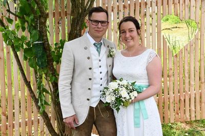 Ein Mann und eine Frau stehen eng nebeneinander vor einem Holzzaun, der Mann trägt einen hellen Anzug und die Frau hält einen Strauß weißer Blumen.