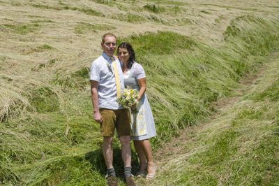 Ein Paar steht in einem Grasfeld, die Frau hält einen Blumenstrauß, beide lächeln in die Kamera.