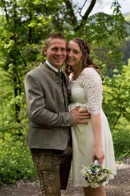 Ein lächelndes Paar in traditioneller Kleidung steht eng beieinander in einem üppigen grünen Wald. Der Mann trägt eine graue Jacke mit Knöpfen, und die Frau hält einen Blumenstrauß.