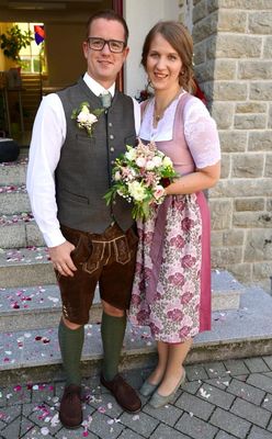 Ein Mann und eine Frau stehen auf einer Treppe und halten einen Blumenstrauß. Der Mann trägt eine Weste und braune Shorts, während die Frau ein Blumenkleid trägt. Sie lächeln beide in die Kamera.