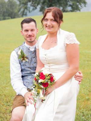 Ein frisch verheiratetes Paar sitzt lächelnd auf einem Grasfeld und hält einen Blumenstrauß. Der Bräutigam trägt eine blaue Weste und die Braut ein weißes Kleid.