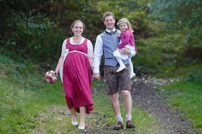 Eine lächelnde schwangere Frau in einem traditionellen Kleid hält Händchen mit einem Mann in traditioneller bayerischer Kleidung, der ein kleines Mädchen auf der Schulter trägt. Sie gehen auf einem Grasweg.