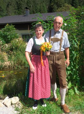 Ein Mann und eine Frau in traditioneller deutscher Kleidung stehen in einem Garten mit einem Teich und einem Haus im Hintergrund.