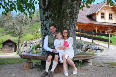 Eine Familie mit drei Personen, ein Mann, eine Frau und ein kleines Mädchen, sitzen auf einer Bank unter einem großen Baum und posieren lächelnd für ein Foto.