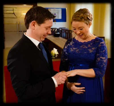 Ein Mann und eine Frau, beide lächelnd, stehen eng beieinander. Der Mann hält die Hand der Frau, die ein blaues Kleid mit einem Blumenbrosche trägt.