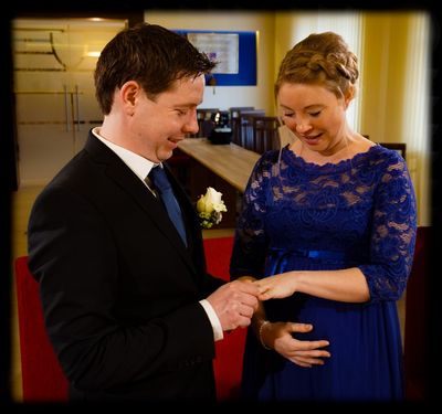 Ein Mann und eine Frau, beide lächelnd, stehen eng beieinander. Der Mann hält die Hand der Frau, die ein blaues Kleid mit einem Blumenbrosche trägt.