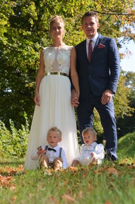 Eine vierköpfige Familie, bestehend aus einer Frau in einem weißen Kleid, einem Mann im Anzug und zwei kleinen Kindern, posiert für ein Foto in einem Park.