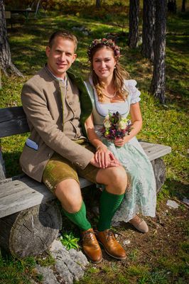 Ein junges Paar in traditioneller deutscher Kleidung sitzt auf einer Holzbank im Park. Die Frau hält einen Blumenstrauß.