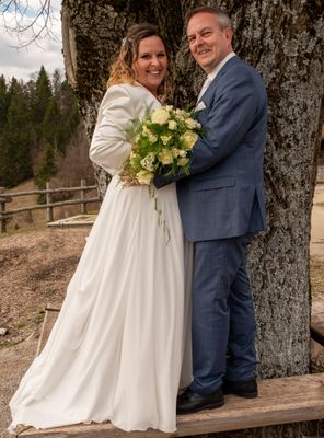 Eine lächelnde Braut in einem weißen Kleid und ein Bräutigam in einem blauen Anzug stehen zusammen, halten einen Blumenstrauß, vor einem Baumstamm.
