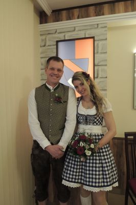 Ein Mann und eine Frau in traditioneller Kleidung stehen lächelnd vor einer dekorativen Wand.
