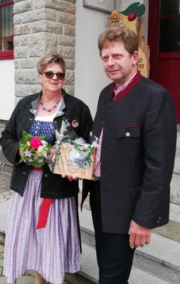 Ein Mann und eine Frau in traditioneller Kleidung stehen zusammen, lächeln und halten einen Blumenstrauß und eine Geschenkbox.