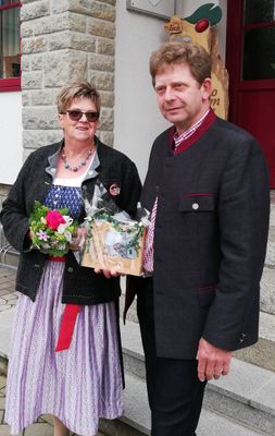 Ein Mann und eine Frau in traditioneller Kleidung stehen zusammen, lächeln und halten einen Blumenstrauß und eine Geschenkbox.
