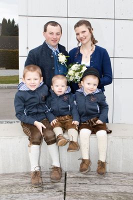 Eine Familie mit vier Personen, darunter zwei Erwachsene und zwei Kinder, gekleidet in traditionelle bayerische Kleidung, posiert für ein Foto. Die Erwachsenen lächeln, und die Kinder sitzen auf einer Betonbank.