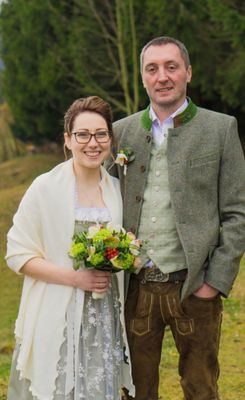 Ein lächelndes Paar, die Frau hält einen Blumenstrauß, steht auf einem grünen Feld. Der Mann trägt traditionelle deutsche Kleidung.